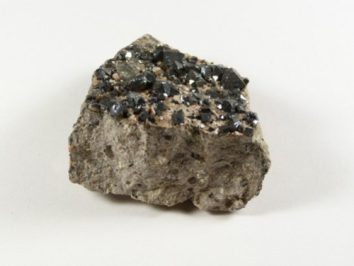 Magnetyt - kamień naturalny, surowy, nieszlifowany. Szara skała z połyskującymi, czarnymi kryształami na powierzchni, na jasnym tle.