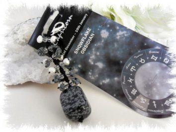 Wisiorek z obsydianu śnieżnego na tle opakowania z motywem astrologicznym.