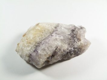 Fluoryt blue john - kamień naturalny, surowy, nieszlifowany. Nieregularny, biało-fioletowy minerał leży na białym tle.