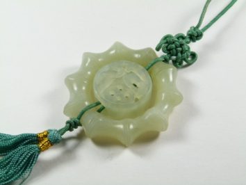 Jasny jadeitowy amulet z zielonym sznurkiem i ozdobnym węzłem leży na białym tle.