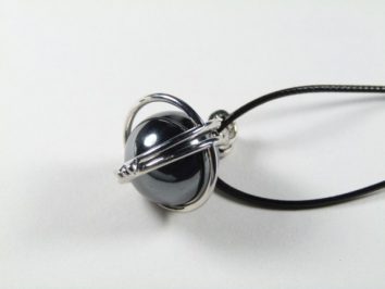 Wisiorek z czarną kulką w srebrnej oprawie na czarnym rzemyku, na białym tle.