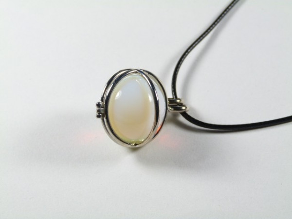 Srebrny wisior z białym, owalnym kamieniem na czarnym sznurku, na jasnym tle.