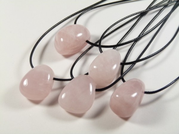Różowy kwarc gładzony z dziurką na sznureczku - wisiorek. Pięć naszyjników z różowego kwarcu na czarnych sznurkach leży na białym tle.