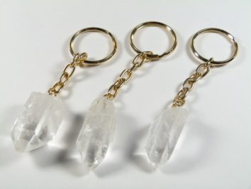 Trzy breloczki z kryształami kwarcu na złotych łańcuszkach, ułożone na białym tle.