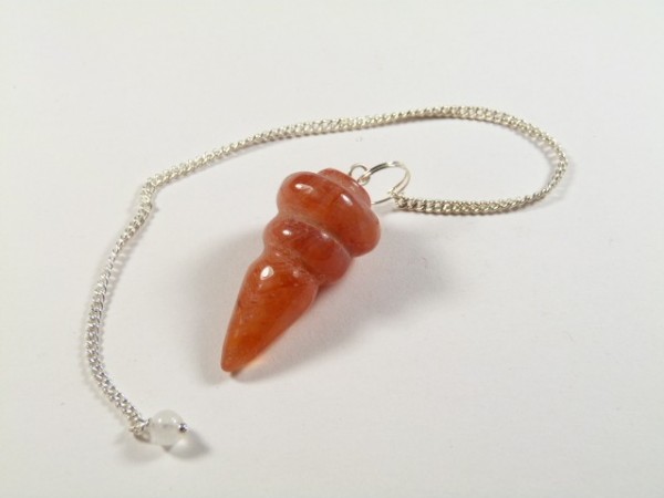 Wahadełko karneol. Wahadło z czerwonego kamienia na srebrnym łańcuszku, leżące na białym tle.