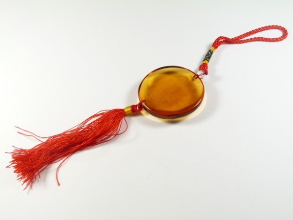 Okrągła zawieszka z bursztynu z czerwoną frędzlą i plecioną rączką na białym tle.