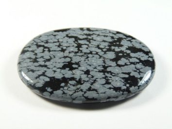 Obsydian śnieżny - kamień gładzony, polerowany, szlifowany. Czarny, płaski kamień z szarymi plamkami przypominającymi śnieg, na białym tle.