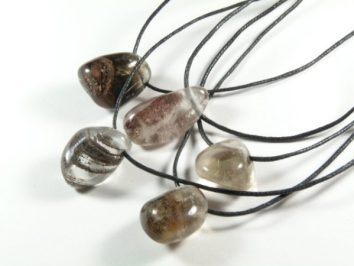 Kryształ górski z inkluzją gładzony z dziurką na sznureczku - wisiorek. Pięć wisiorków z kamieniami na czarnych sznurkach leży na białym tle.