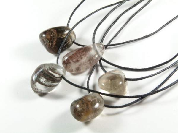 Kryształ górski z inkluzją gładzony z dziurką na sznureczku - wisiorek. Pięć wisiorków z kamieniami na czarnych sznurkach leży na białym tle.