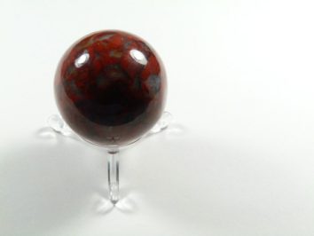 Kula jaspis makowy, średnica 4,5 cm. Czerwona kula z kamienia ozdobnego na przezroczystej podstawce, na białym tle.