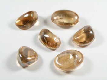 Kwarc tangerine aura - kamień gładzony, polerowany, szlifowany i bębnowany w formie otoczaka. Sześć polerowanych, jasnobrązowych kamieni leży na białym tle.
