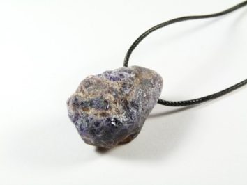 Surowy fioletowy kamień na czarnym sznurku, leżący na białym tle.