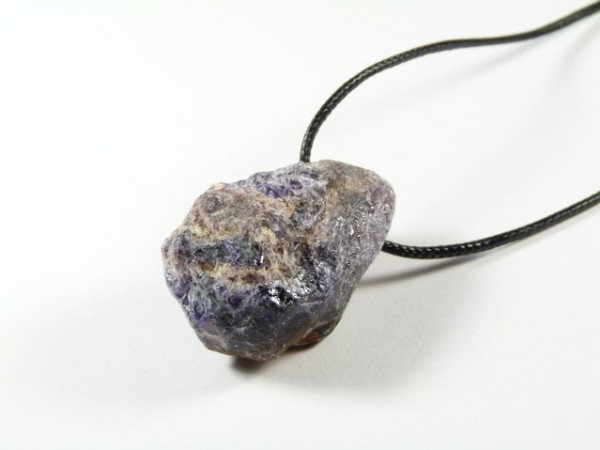Surowy fioletowy kamień na czarnym sznurku, leżący na białym tle.