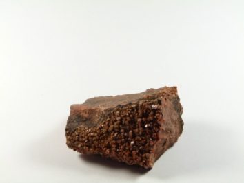 Wanadynit - kamień naturalny, surowy, nieszlifowany. Brązowy kamień z widocznymi kryształkami na jednej z powierzchni, na białym tle.
