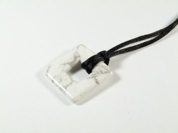 Kwadratowy biały wisiorek z żyłkowanego kamienia na czarnym sznurku leży na białym tle.