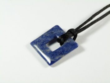 Niebieski, kwadratowy wisiorek z kamienia na czarnym sznurku, leżący na białym tle.