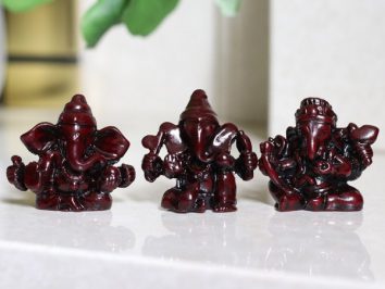 Trzy małe figurki Ganeshy ustawione obok siebie na jasnej powierzchni.