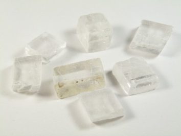 Spar islandzki - kamień naturalny, surowy, nieszlifowany. Siedem przezroczystych kryształów leży na białym tle.