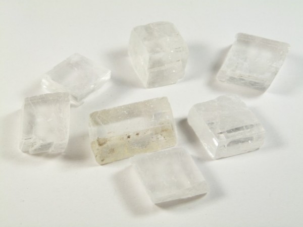 Spar islandzki - kamień naturalny, surowy, nieszlifowany. Siedem przezroczystych kryształów leży na białym tle.