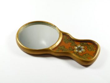 Małe lusterko z ozdobną rączką w kwiatowy wzór leżące na białym tle.