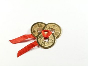 Trzy chińskie monety z otworem połączone czerwoną wstążką na białym tle.