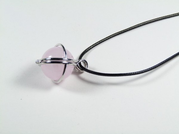 Wisior z różowym kamieniem w metalowej oprawie na czarnym sznurku, leży na białym tle.