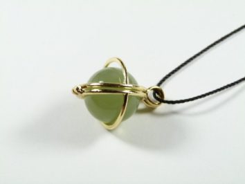 Wisiorek z zieloną kulką otoczoną złotymi obręczami na czarnym sznurku, na białym tle.