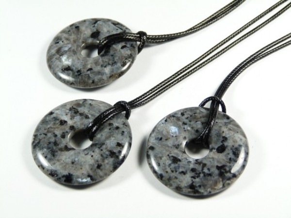 Trzy okrągłe wisiorki z szarego kamienia na czarnych sznurkach leżą na białym tle.