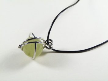 Naszyjnik z zielonym kamieniem oprawionym w metal, na czarnym sznurku, na jasnym tle.