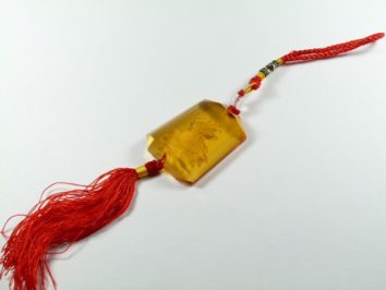 Bursztynowy amulet z czerwonym frędzlem i sznurkiem leży na białym tle.