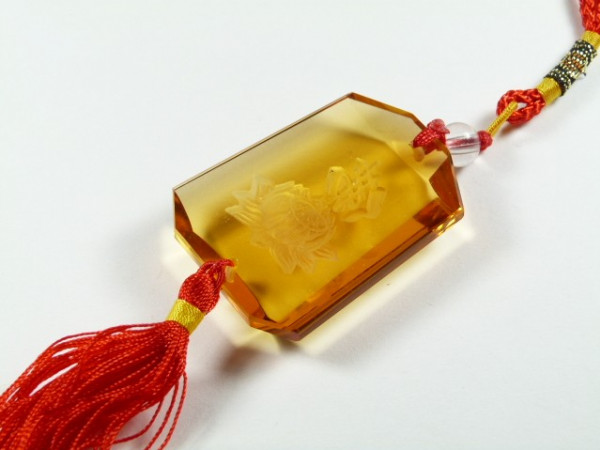 Bursztynowy wisiorek z kwiatowym grawerem, ozdobiony czerwonym sznurkiem i frędzlami.