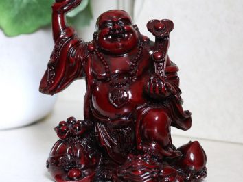 Ciemnoczerwona figurka uśmiechniętego Buddy z uniesioną ręką, siedzącego na podstawce.