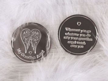 Dwa srebrne medaliki z motywem anielskich skrzydeł, leżące na białych piórkach.