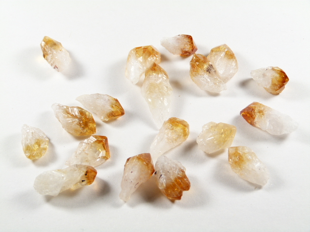 Cytryn - kamień naturalny surowy nieszlifowany. Na zdjęciu widać kilkanaście naturalnych kryształków cytrynu o złocisto-bursztynowych końcówkach i przejrzystej strukturze, ułożonych na białym tle. Cytryn kamień bogactwa.