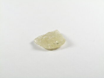 Na zdjęciu znajduje się naturalny kryształ hiddenitu zielonego w odcieniu jasnozielonym, o surowej, lekko przejrzystej strukturze, leżący na białym tle.