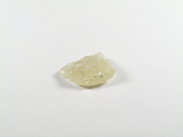 Na zdjęciu znajduje się naturalny kryształ hiddenitu zielonego w odcieniu jasnozielonym, o surowej, lekko przejrzystej strukturze, leżący na białym tle.