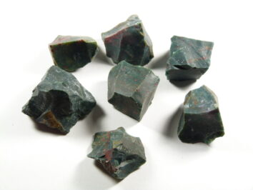 Bloodstone właściwości – surowe kamienie heliotropu. Siedem surowych, zielonych kamieni z czerwonymi plamkami na białym tle.