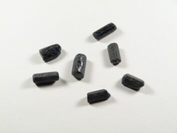 Czarny turmalin kamień ochronny w formie surowych, podłużnych kryształów na jasnym tle. Siedem czarnych, cylindrycznych pastylek leży na białym tle.