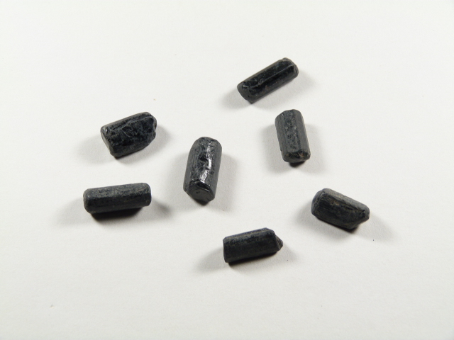 Czarny turmalin kamień ochronny w formie surowych, podłużnych kryształów na jasnym tle. Siedem czarnych, cylindrycznych pastylek leży na białym tle.