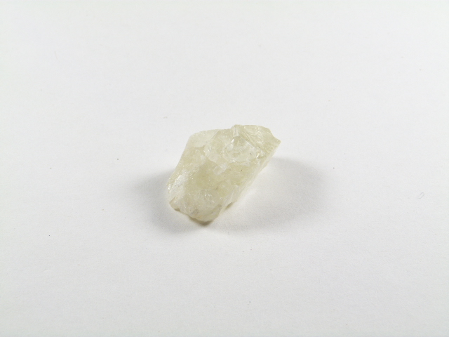 Kamień hiddenit zielony w formie surowego, jasnego kryształu o bardzo delikatnym seledynowym odcieniu. Nieoszlifowany, prawie biały kryształ soli leży na białym tle.