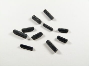 Kamień czarny turmalin w formie małych, surowych, nieszlifowanych kamyczków. Dziesięć czarnych, cylindrycznych granulek leży na białej powierzchni.