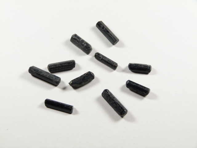 Kamień czarny turmalin w formie małych, surowych, nieszlifowanych kamyczków. Dziesięć czarnych, cylindrycznych granulek leży na białej powierzchni.